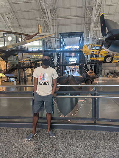 Sidney Boakye wearing a NASA shirt and a face mask in front of antique planes. 