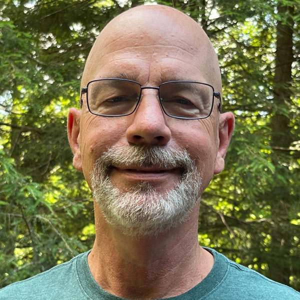Charles White wears a sage-green t-shirt and glasses in a wooded area for his profile in the Department of Mechanical Engineering.