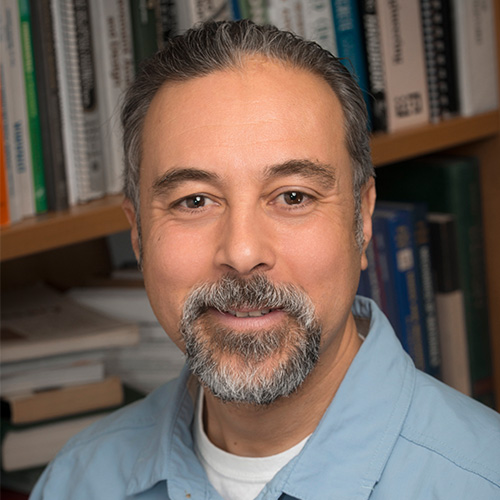 Mason associate professor Burak Tanyu wears a blue collared shirt and stands in a library for his faculty profile