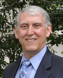 This headshot of Art Pyster in front of a tree and wearing a blue shirt and tie with a black suit jacket for his faculty profile. 
