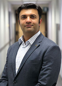 Ali Raz stands in an office hallway wearing a dark-gray suit and light shirt in his faculty profile for Mason's Systems Engineering and Operations Research department