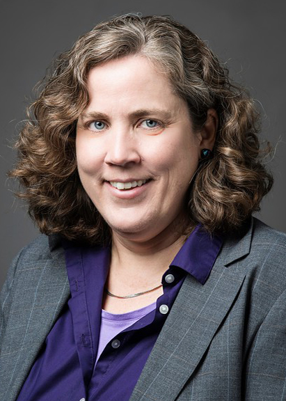 This is a headshot of Kathleen Wage in a purple shirt and gray suit for her faculty profile at George Mason University.