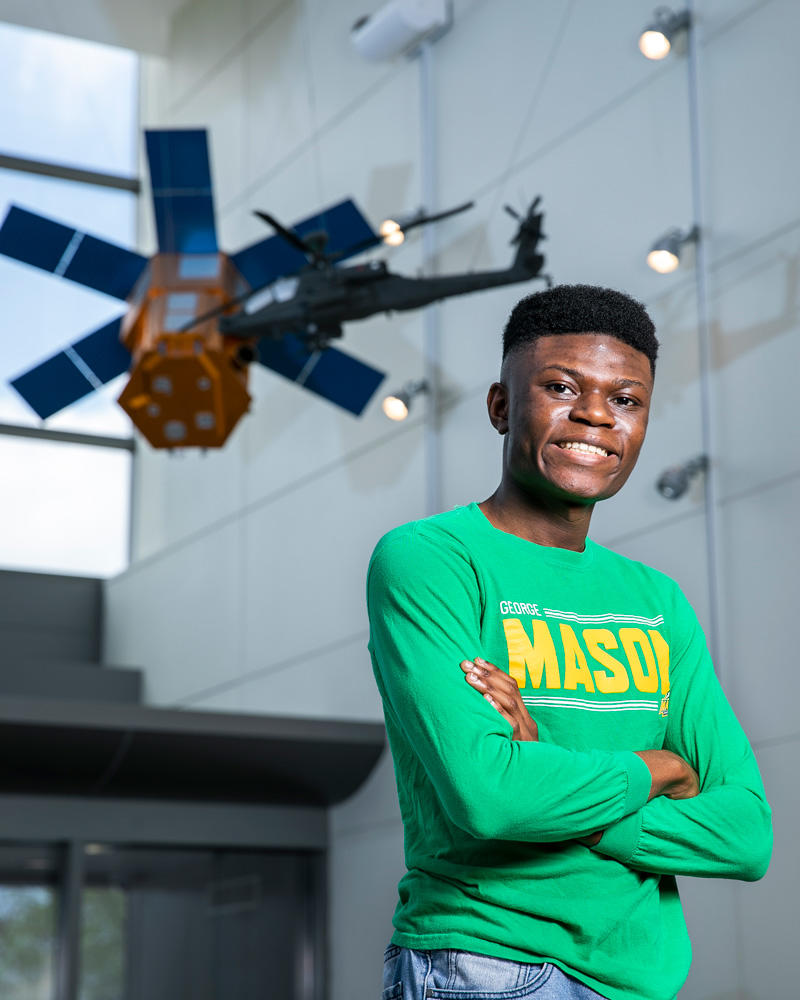 Sidney Boakye stands in the gallery of satellites at the U.S. Air and space museum. He is wearing a green George Mason University t-shirt. He is smiling and proud. 