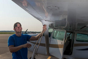 White young adult male wears a baseball cap and stands next to a plane on a tarmac